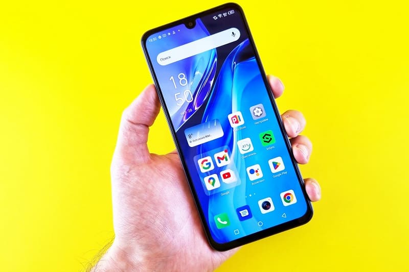 Uma mão segurando um smartphone mostrando a tela principal cheia de ícones. Ao fundo, um plano amarelo