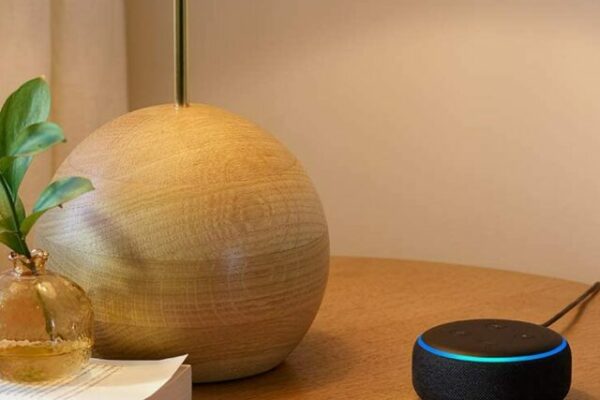 Dispositivo Amazon Echo Dot 3 com Alexa, em cima de uma mesa de cor madeira e ao lado de um abajur.