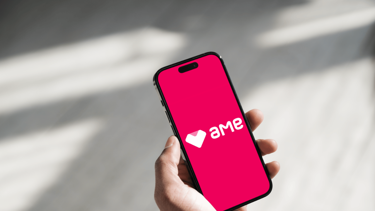 Uma mão segurando um smartphone mostrando o logotipo da Ame Digital em um fundo rosa pink. Imagem: ilustração Panorama Tecnológico.