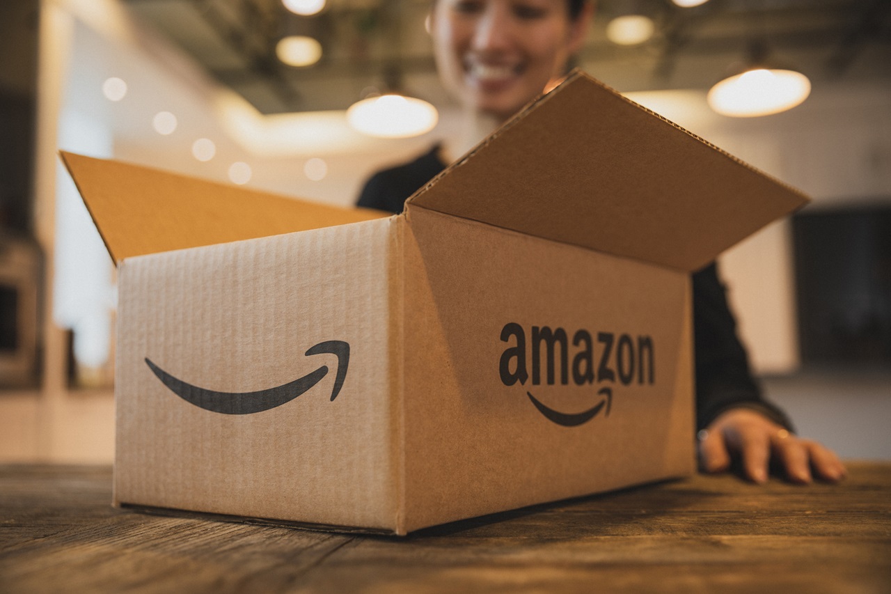 Uma caixa de papelão da Amazon aberta sobre uma superfície de madeira. O icônico logotipo de sorriso da Amazon é visível na lateral da caixa. Ao fundo, um ambiente interno levemente desfocado, com uma pessoa sentada à mesa ou escrivaninha, cuja face está obscurecida por um bloco de anonimização. Imagem: Amazon