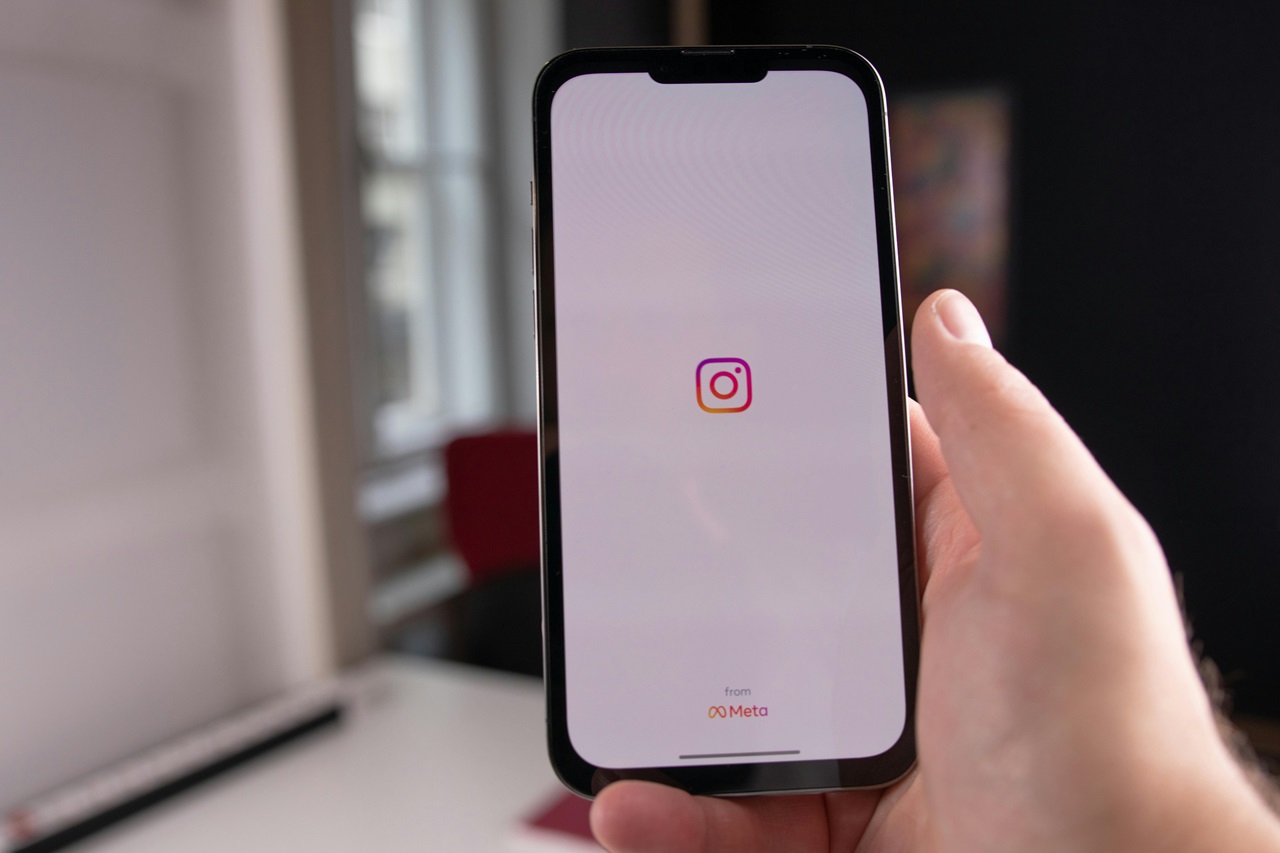 A imagem mostra uma mão segurando um smartphone com a tela visível para o espectador. O smartphone exibe o logotipo do Instagram, que é um ícone de câmera, no centro de um fundo branco com uma barra superior em gradiente rosa e laranja. Abaixo do logotipo, há um texto que diz “from META”. Foto de DlxMedia.hu/Pexels
