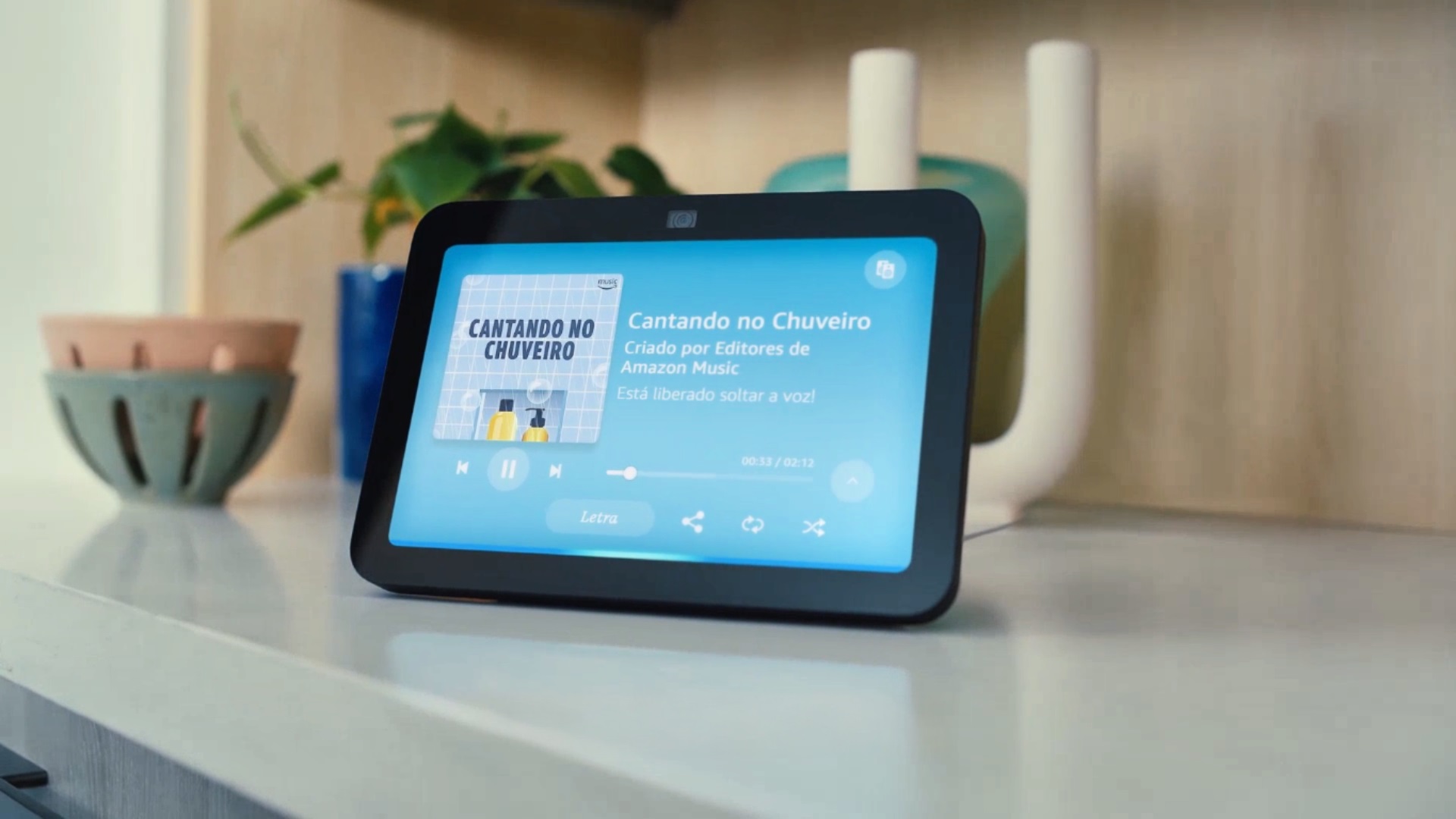 A imagem mostra um dispositivo Echo Show 8 da Amazon em uma bancada. A tela do dispositivo exibe a interface de reprodução de música, com a capa do álbum