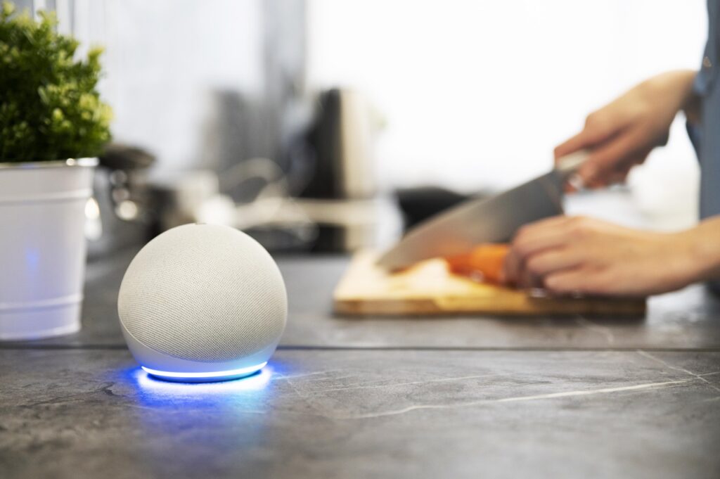 A imagem mostra uma as mãos de uma pessoa cortando legumes em cima de uma bancada de uma cozinha. A imagem está um pouco desfocada e mostra um vaso de planta e um dispositivo Echo Dot 5ª geração com Alexa na cor branca. Imagem de Freepik.