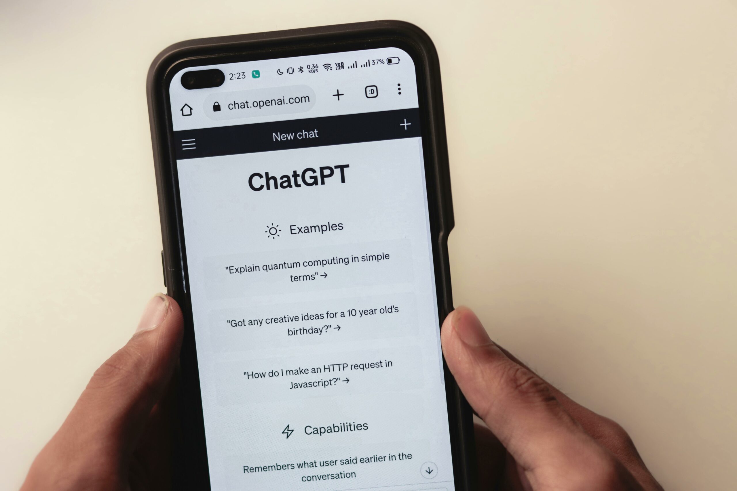 Imagem mostrando uma pessoa segurando um smartphone que exibe a interface do ChatGPT no site chat.openai.com.A tela do telefone mostra a seção 'Examples' com as seguintes perguntas: 'Explain quantum computing in simple terms,' 'Got any creative ideas for a 10 year old's birthday?,' e 'How do I make an HTTP request in Javascript?.' A seção 'Capabilities' também é visível parcialmente na parte inferior da tela. Imagem: Sanket Mishra / Pexels.
