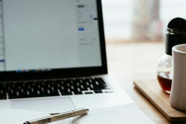 Imagem mostrando uma bancada de trabalho sobre uma mesa de madeira. Há um laptop aberto exibindo um documento na tela, com uma página de rede social parcialmente visível atrás. Na frente do laptop, há um caderno aberto com uma caneta sobre ele. À direita do caderno, há uma caneca branca em um porta-copos de madeira, um pote de vidro com um pequeno buquê de flores e um pequeno recipiente de vidro com um líquido escuro. A cena está posicionada perto de uma janela, proporcionando uma vista desfocada do lado de fora. Créditos: Nick Morrison / Unsplash