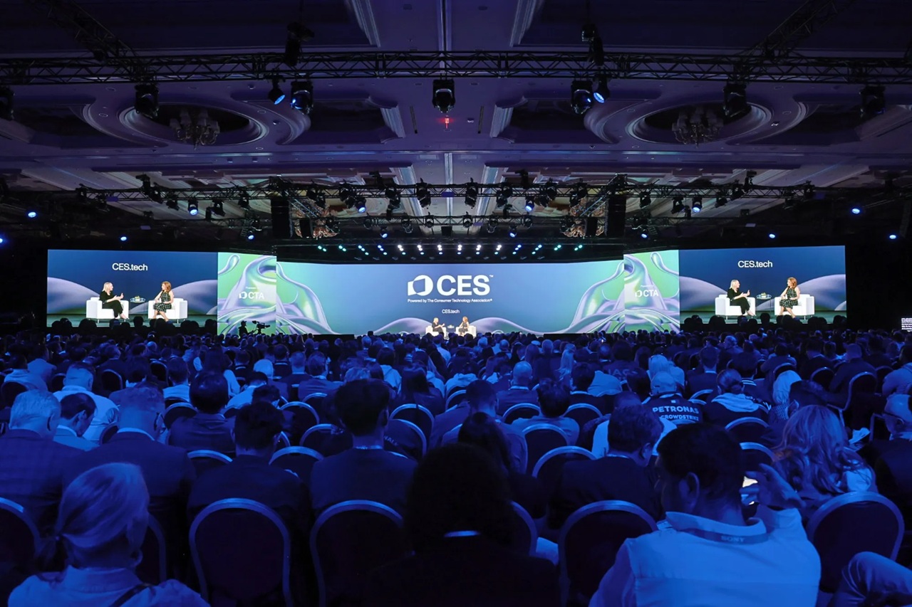 Imagem de um grande evento ou conferência com um palco e uma plateia sentada. No palco, há uma grande tela exibindo o logotipo do CES (Consumer Electronics Show), um dos maiores eventos de tecnologia do mundo. Duas pessoas estão sentadas no palco, provavelmente participando de uma discussão ou entrevista. A plateia, composta por uma diversidade de pessoas, está atenta, assistindo à apresentação. Imagem: divulgação CES.