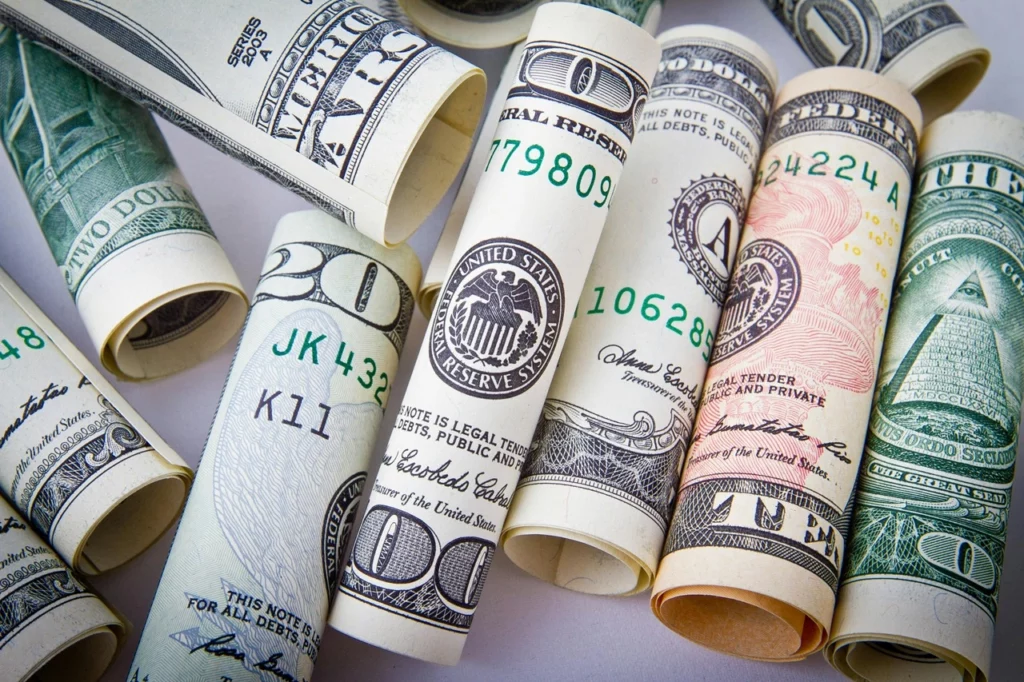 Uma imagem em close-up de várias notas de dólar americano enroladas de diferentes denominações, incluindo $20, $10 e $5, dispostas de maneira espalhada sobre uma superfície branca. As notas exibem partes diferentes de seus designs, como o selo do Federal Reserve, números de série e outros elementos distintivos do papel-moeda dos EUA. A imagem destaca a diversidade e os detalhes das cédulas americanas. Créditos: Pixabay / Pexels.