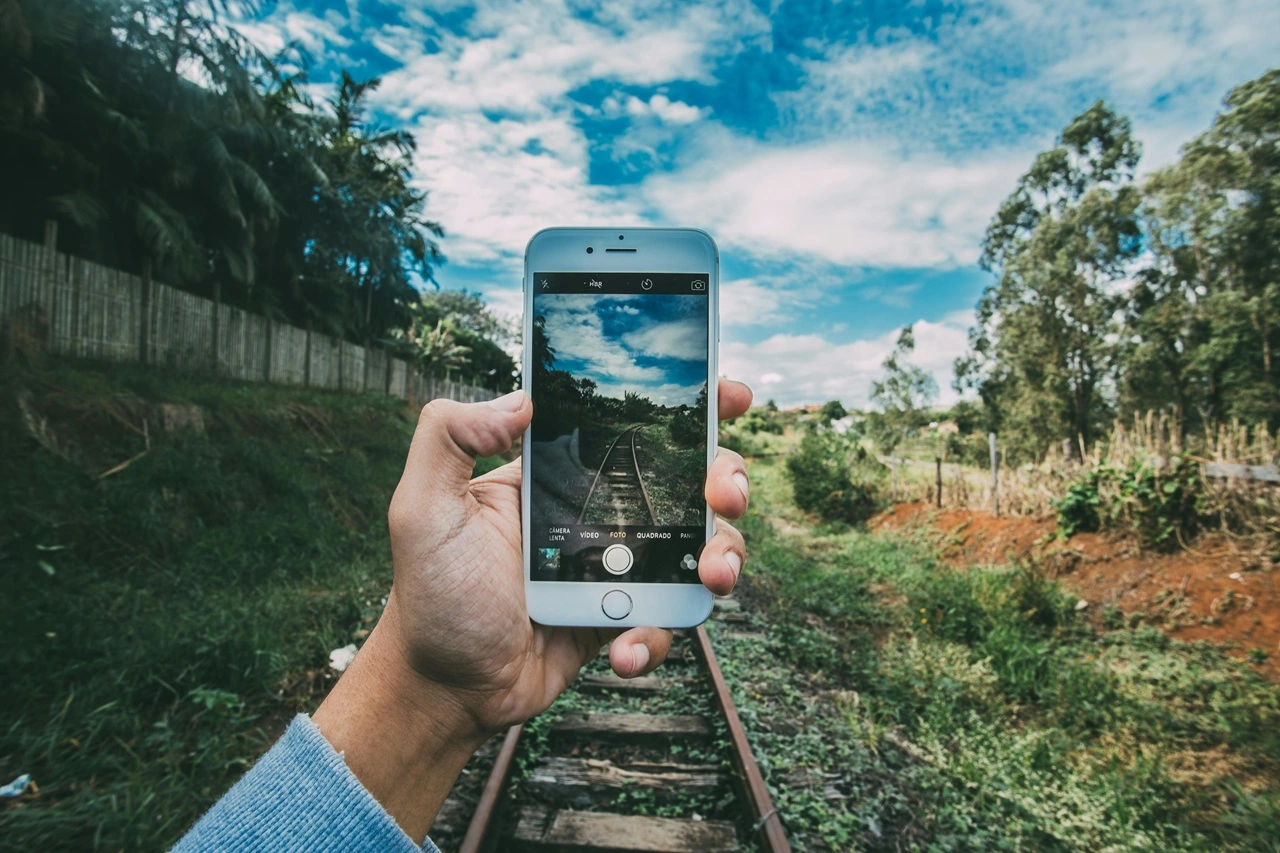 A imagem mostra uma mão segurando um smartphone, capturando uma foto de uma trilha ferroviária que se estende à distância. A tela do smartphone exibe a mesma trilha ferroviária, criando um efeito em camadas onde a cena real e a captura digital estão alinhadas. O fundo inclui vegetação, árvores e um céu parcialmente nublado. Esta imagem é interessante porque destaca a convergência entre a realidade e a representação digital, mostrando como a tecnologia pode capturar e refletir o mundo ao nosso redor. Créditos: Kaique Rocha / Pexels.