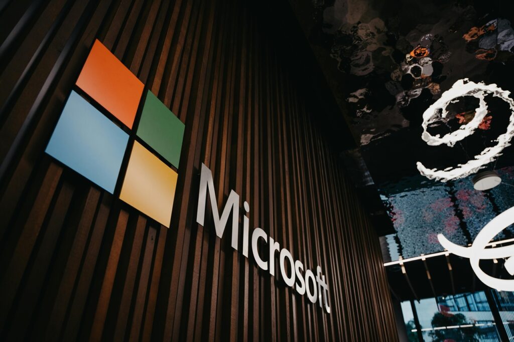 Imagem que mostra o logotipo e o nome da empresa Microsoft, fixados em uma parede ripada de cor de madeira escura. Créditos: Angel Bena / Pexeis.