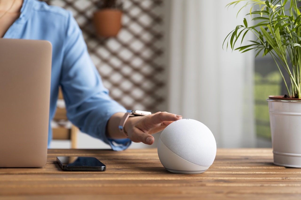 A imagem mostra uma pessoa interagindo com um dispositivo inteligente de formato esférico, que está sobre uma mesa de madeira. A pessoa está usando uma camisa azul e um relógio de pulso, e há um laptop e um smartphone também sobre a mesa. Ao fundo, há uma planta em um vaso branco e uma parede com um padrão de treliça. A cena sugere um ambiente de trabalho ou estudo, onde a tecnologia é utilizada para facilitar tarefas diárias. Créditos: Freepik.