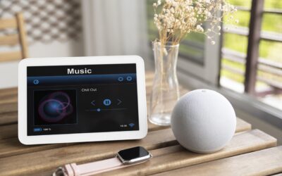 A imagem mostra uma mesa de madeira com um dispositivo inteligente de tela branca exibindo uma interface de música com a faixa 