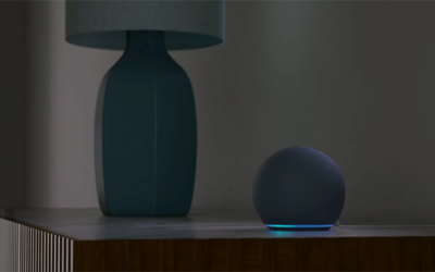 A imagem apresenta um dispositivo Echo Dot com Alexa, com formato esférico e uma base iluminada por luz azul, posicionado sobre uma superfície de madeira. Ao lado, há um abajur com base azul e cúpula cinza. O fundo da imagem está desfocado, destacando os objetos principais. Créditos: Amazon divulgação.