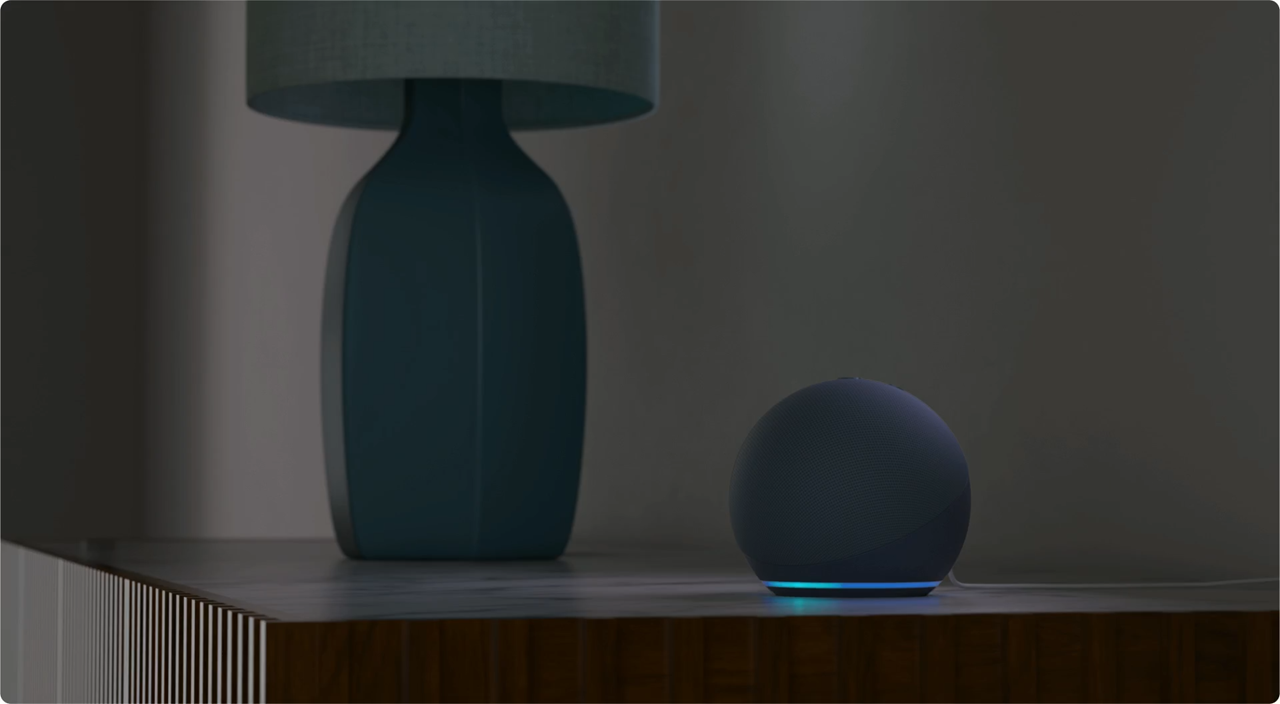 A imagem apresenta um dispositivo Echo Dot com Alexa, com formato esférico e uma base iluminada por luz azul, posicionado sobre uma superfície de madeira. Ao lado, há um abajur com base azul e cúpula cinza. O fundo da imagem está desfocado, destacando os objetos principais. Créditos: Amazon divulgação.