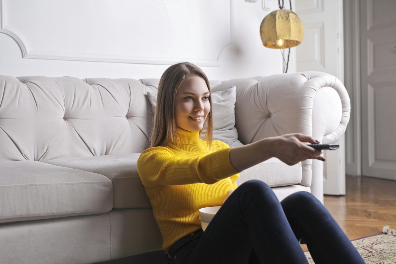 A imagem mostra uma mulher loira sentada no chão da sala encostada em um sofá branco. Ela está de calça com uma blusa amarela segurando um controle remoto apontando para frente. A imagem sugere que ela esteja comandando uma TV. Créditos: Andrea Piacquadio / Pexels.