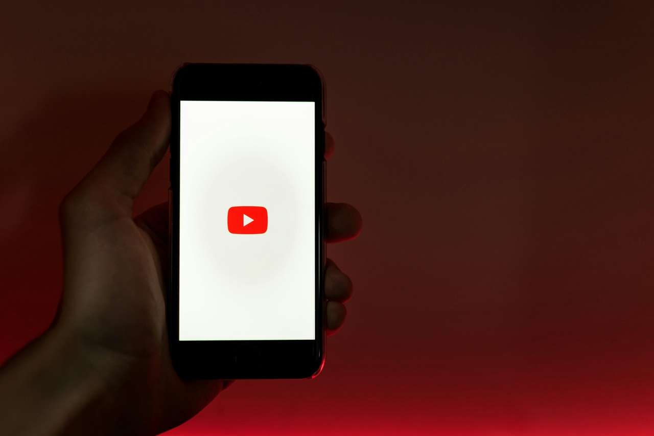 Uma mão segurando um smartphone cuja tela exibe o logotipo do YouTube, caracterizado por um triângulo branco dentro de um retângulo vermelho. O fundo da imagem é escuro, com um tom avermelhado mais intenso na parte inferior. Créditos: Szabó Viktor / Pexels.