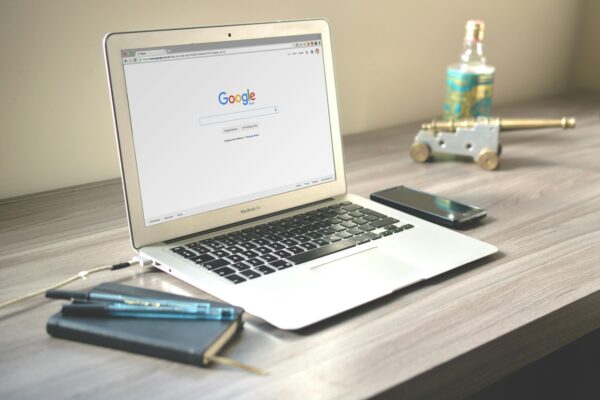 Google volta a exibir cotação do dólar na busca após correções Notebook MacBook Air sobre uma mesa de madeira exibindo a página inicial do Google Brasil. A tela mostra a barra de pesquisa do Google com as opções