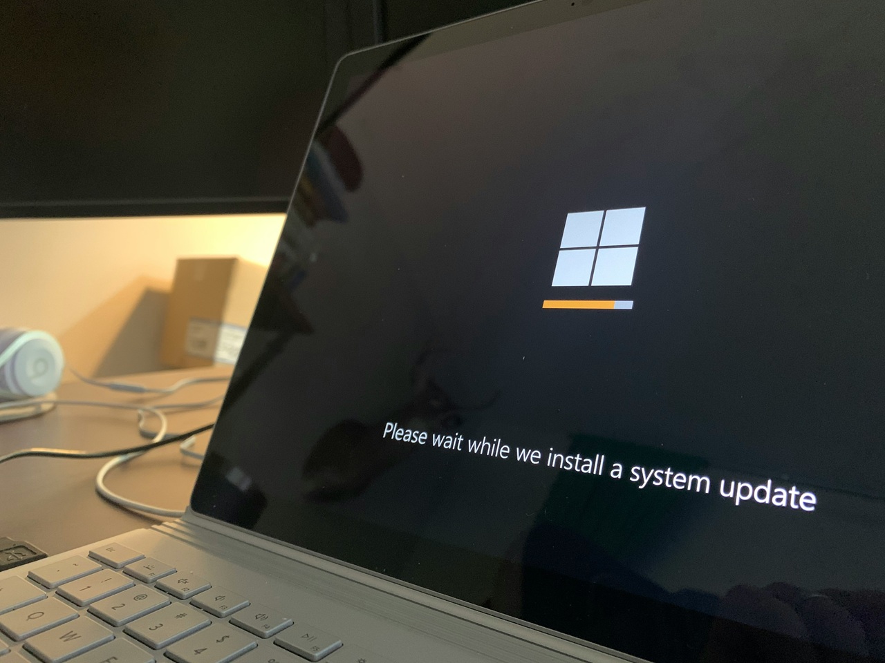 Imagem de um laptop com a tela exibindo uma mensagem de atualização do sistema operacional Windows. A mensagem na tela diz: 'Please wait while we install a system update'. A barra de progresso está parcialmente preenchida, indicando que a atualização está em andamento. No fundo, há uma mesa com alguns objetos, incluindo um par de fones de ouvido e uma caixa. Créditos: imagem de Clint Petterson / Unsplash