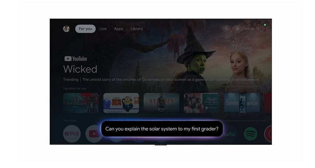 A tela de uma TV exibe a interface do Google TV, destacando um vídeo do YouTube chamado 'Wicked' com uma descrição sobre as bruxas de Oz. A seção 'Para você' apresenta recomendações de vídeos e aplicativos como Netflix, Prime Video, Hulu e Spotify. No centro da tela, há uma barra de pesquisa com a pergunta: 'Can you explain the solar system to my first grader?' (Você pode explicar o sistema solar para meu aluno da primeira série?). Créditos: Google/divulgação.