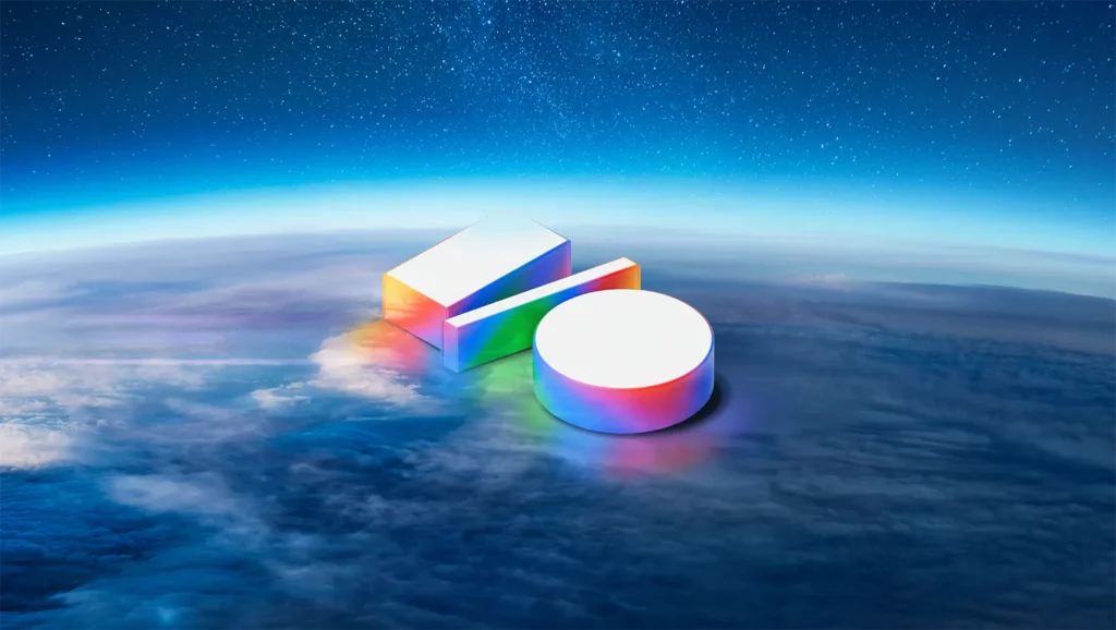 A imagem mostra uma representação estilizada do logotipo do Google I/O, que consiste em duas formas geométricas tridimensionais: um cilindro e um prisma retangular, ambos com um efeito de arco-íris. As formas estão posicionadas sobre uma vista da Terra a partir do espaço, com uma camada de nuvens e um céu estrelado ao fundo. Créditos: Google.