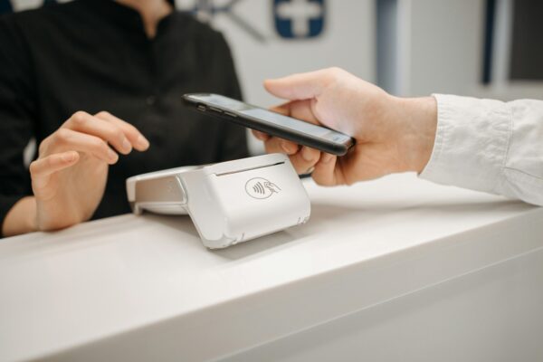 Aplicativo Nubank ganha função de Pix por aproximação A imagem mostra uma pessoa segurando um smartphone próximo a um terminal de pagamento sem contato, sugerindo uma transação de pagamento digital. A pessoa está vestida com uma camisa branca e o terminal de pagamento é branco com um símbolo de pagamento sem contato. Outra pessoa, vestida de preto, está ao fundo, possivelmente um atendente. Créditos: imagem de Pavel Danilyuk / Pexels.