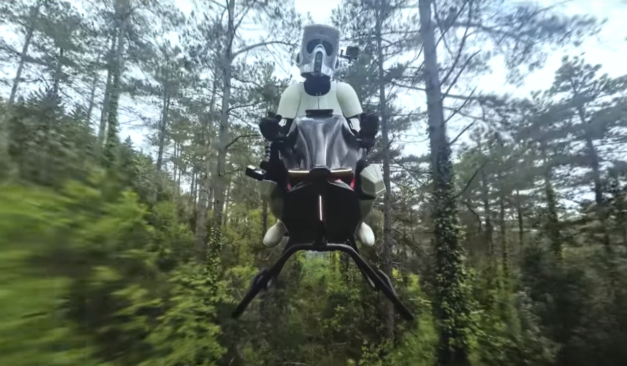 Um Stormtrooper de Star Wars pilota uma moto voadora futurista em uma floresta densa, cercado por árvores altas e vegetação exuberante. A moto exibe luzes na frente e parece flutuar no ar, criando um contraste marcante entre tecnologia e natureza. Créditos: Volonaut.