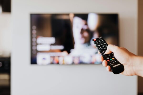 Como criar um ambiente de cinema usando Fire TV e Echo Studio A imagem mostra uma mão segurando um controle remoto da marca Samsung, apontando para uma televisão. A tela da TV exibe uma interface de streaming com várias opções de conteúdo, incluindo uma imagem desfocada de uma pessoa. A cena sugere que alguém está prestes a selecionar ou mudar o conteúdo na TV usando o controle remoto. Créditos: imagem de Kaboompics.com / Pexels.