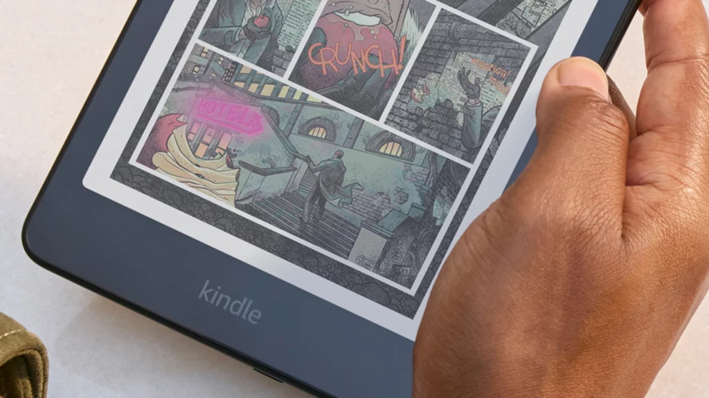 Mão segurando um dispositivo Kindle exibindo uma página de quadrinhos colorida. A tela mostra cenas vibrantes como uma pessoa mordendo uma maçã com o som ‘CRUNCH!’, um letreiro de hotel em neon rosa, e uma escadaria urbana. A imagem destaca o uso do Kindle para leitura de quadrinhos com riqueza de detalhes e cores. Créditos: divulgação Amazon.