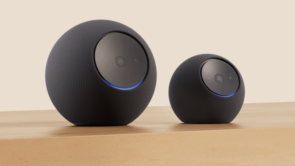 Dois alto-falantes inteligentes da Amazon estão posicionados sobre uma superfície de madeira. À direita, o Echo Dot Max, menor e compacto, com acabamento escuro e painel de controle circular com botões sensíveis ao toque. À esquerda, o Echo Studio, maior e mais robusto, com design semelhante e luz azul ao redor do painel, indicando que está ligado. Ambos os dispositivos apresentam ícones de volume e microfone, destacando suas funções de assistente de voz. A imagem transmite modernidade e sofisticação em tecnologia doméstica. Créditos: Amazon divulgação.