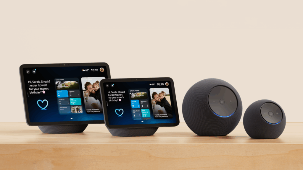 Conjunto dos novos dispositivos inteligentes da Amazon exibidos sobre uma superfície de madeira com fundo neutro. Inclui os modelos Echo Dot Max, Echo Studio (2ª geração), Echo Show 8 (4ª geração) e Echo Show 11, todos com design moderno e integração com Alexa+. Os displays inteligentes mostram informações personalizadas como previsão do tempo, eventos do calendário e uma mensagem interativa, enquanto os alto-falantes esféricos exibem um anel azul iluminado indicando atividade. Créditos: Amazon divulgação.