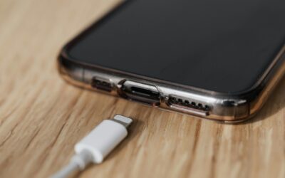 Close-up de um smartphone apoiado sobre uma mesa de madeira, com destaque para a entrada de carregamento na parte inferior e um cabo Lightning branco próximo, prestes a ser conectado ao aparelho. Créditos: kaboompics / Pexels.