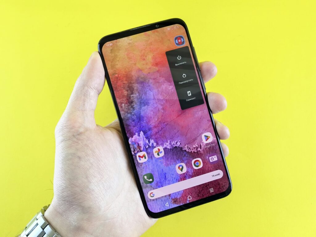 Mão segurando um smartphone sobre fundo amarelo, com a tela inicial visível exibindo ícones de aplicativos como Google Play e outros apps, representando o uso cotidiano de celular e navegação entre aplicativos. Créditos: Andrey Matveev / Pexels.