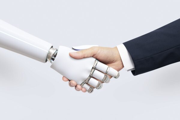 Mão humana apertando a mão de um robô humanoide em uma imagem conceitual sobre inteligência artificial, automação e colaboração entre humanos e máquinas. O fundo claro destaca o contraste entre o braço mecânico branco e a pessoa vestindo terno social. Créditos: rawpixel.com / Freepik