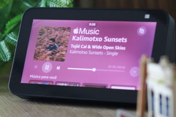 Smart display da linha Amazon Alexa exibindo a interface de reprodução de música, com capa do álbum, controles de mídia e informações da faixa na tela. O dispositivo está apoiado sobre uma mesa em ambiente interno, cercado por elementos decorativos e plantas ao fundo. Créditos: Thiago Japyassu / Pexels.