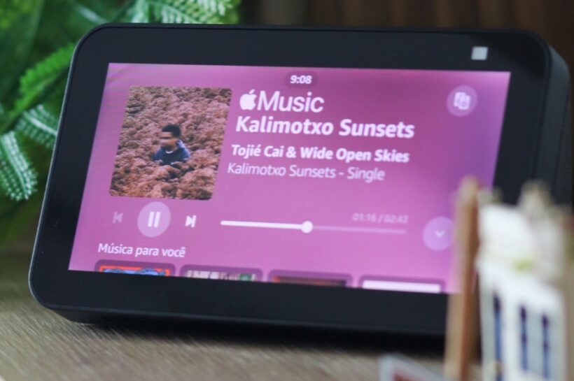 Smart display da linha Amazon Alexa exibindo a interface de reprodução de música, com capa do álbum, controles de mídia e informações da faixa na tela. O dispositivo está apoiado sobre uma mesa em ambiente interno, cercado por elementos decorativos e plantas ao fundo. Créditos: Thiago Japyassu / Pexels.