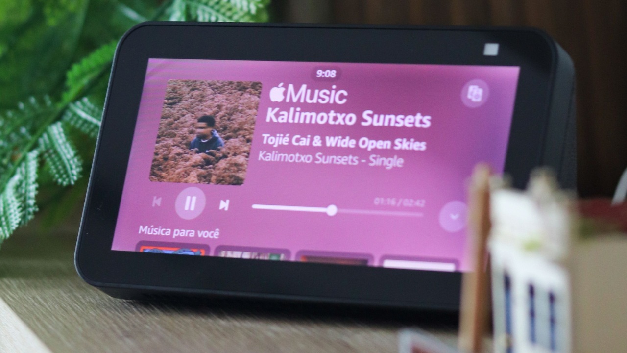 Smart display da linha Amazon Alexa exibindo a interface de reprodução de música, com capa do álbum, controles de mídia e informações da faixa na tela. O dispositivo está apoiado sobre uma mesa em ambiente interno, cercado por elementos decorativos e plantas ao fundo. Créditos: Thiago Japyassu / Pexels.