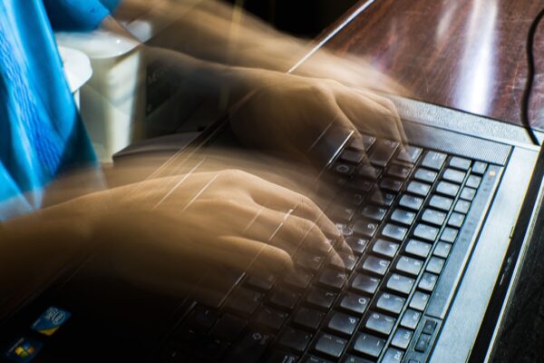 Mãos digitando rapidamente em um notebook, com efeito de movimento que representa atividades de informática, programação, cibersegurança ou ataques virtuais, ilustrando temas relacionados a golpes digitais, hackers e segurança da informação. Créditos: imagem de Nothing Ahead / Pexels.