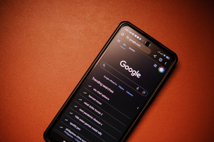 Smartphone exibindo a página inicial do Google em modo escuro no navegador, com a barra de pesquisa e a lista de pesquisas em alta na tela. O aparelho está apoiado sobre uma superfície marrom, em um enquadramento que destaca o uso do mecanismo de busca em dispositivos móveis. Créditos: Click Jeth / Pexels.