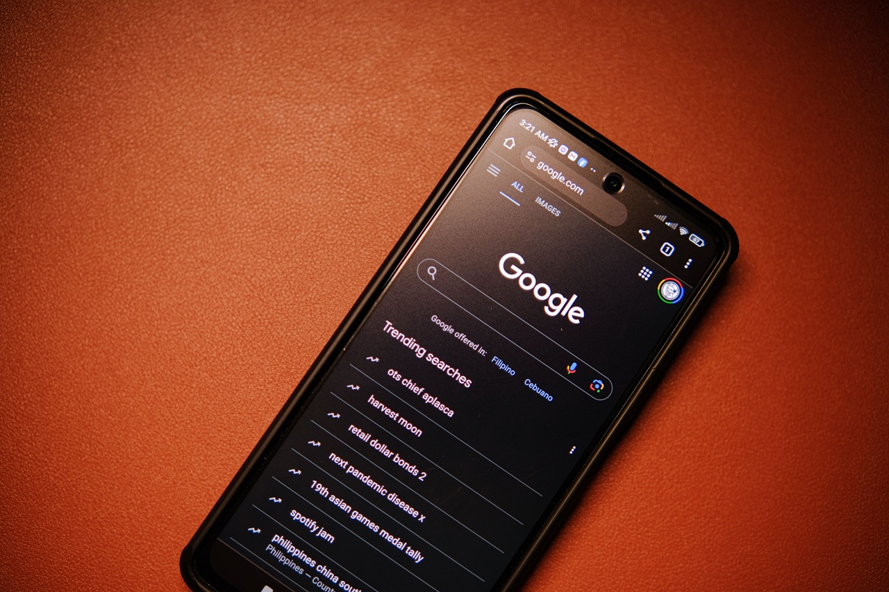 Smartphone exibindo a página inicial do Google em modo escuro no navegador, com a barra de pesquisa e a lista de pesquisas em alta na tela. O aparelho está apoiado sobre uma superfície marrom, em um enquadramento que destaca o uso do mecanismo de busca em dispositivos móveis. Créditos: Click Jeth / Pexels.