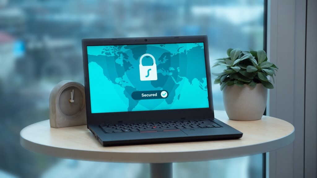 Notebook exibindo uma tela com símbolo de cadeado e indicação de conexão segura, representando proteção de dados e segurança digital. Ao fundo, há uma planta sobre uma mesa próxima a uma janela. Créditos: Dan Nelson / Pexels.
