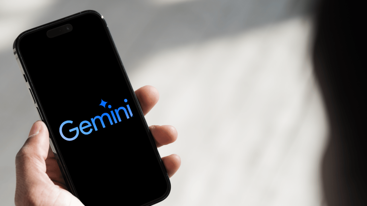 Mão segurando um smartphone com a tela preta exibindo o logotipo azul do Google Gemini, assistente de inteligência artificial do Google.